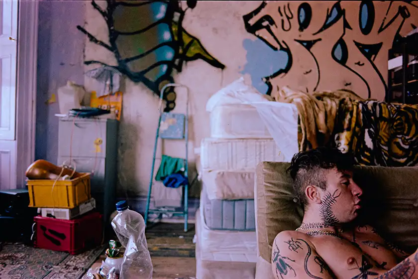 Barry Keoghan sits on a couch shirtless in a messy home in a still from the film Bird, whose actors Nykiya Adams and Jason Buda we interview in this article