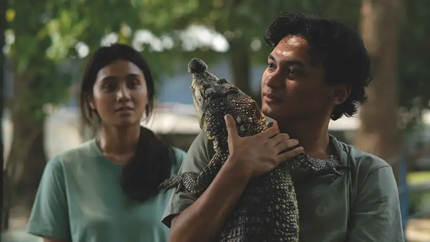 A man holds a crocodile with a woman standing behind him in the film Crocodile Tears