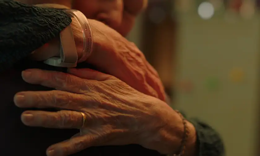 Two elderly hands are shown, belonging to two people who are hugging each other, in a still from the film Familiar Touch