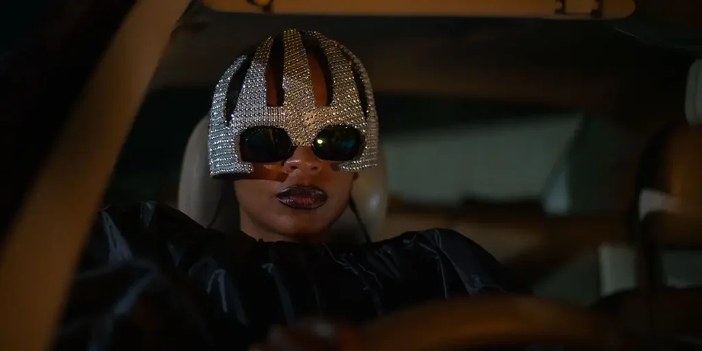 Susan Chardy, as Schula, drives a car at night, wearing sunglasses and a sparkly hat but with a stern expression, in On Becoming a Guinea Fowl