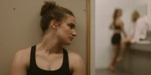 A long woman hides behind a bathroom stall looking at two other girls in a still from the film Julie Keeps Quiet