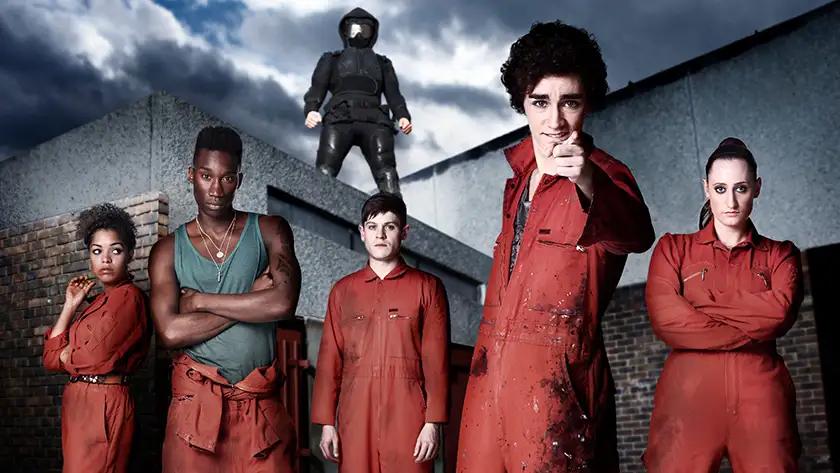 Five teenagers wear orange community service suits looking at the camera in the series Misfits, with a black figure looming over them in the background