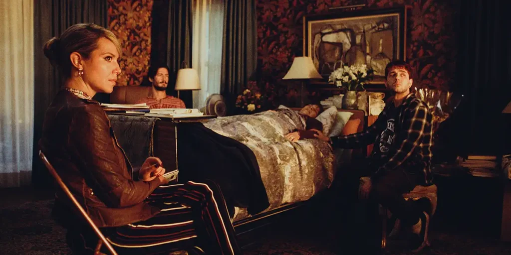 Two people sit next to a bed with someone in it in a still from The Night Logan Woke Up