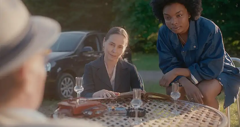 A woman sits by a wooden table in the garden and another woman leans on it, and they both look at a man, in a still from the film Portraits of Dangerous Women