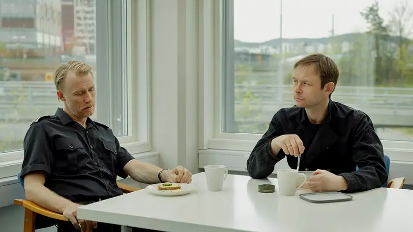 Thorbjørn Harr as “Supervisor” and Jan Gunnar Røise as “Chimney Sweeper” sit at a table drinking coffee and talking in a still from the film Sex, whose director, Dag Johan Haugerud, we interview