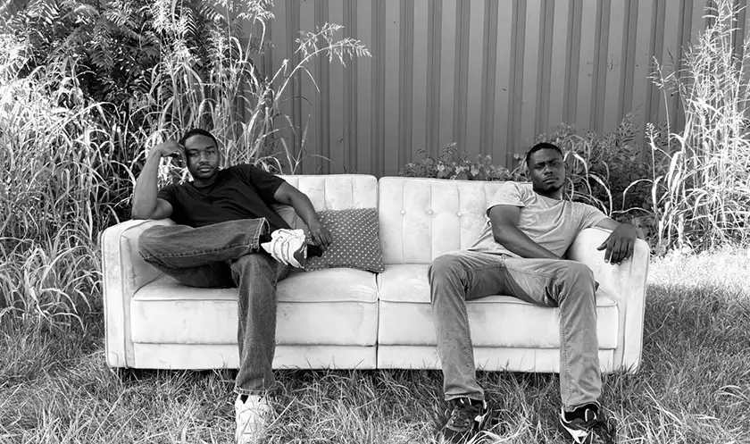 Two men sit on a white couch outdoors in a black and white still from the film Sofa, So Good