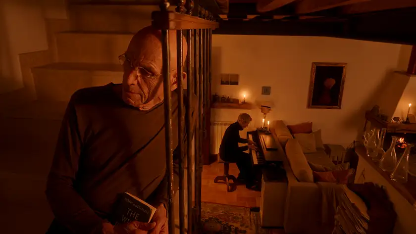 A character stands near a staircase in a house while another character plays the piano in the film Two Strangers Trying Not to Kill Each Other