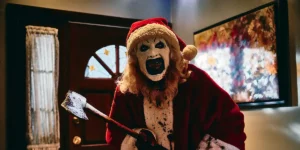 Art The Clown smiles holding an axe and wearing a santa suit and hat in the movie in Terrifier 3