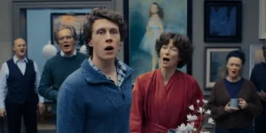 George Mackay and Tilda Swinton sing facing the camera, with some people behind them, in a still from the film The End