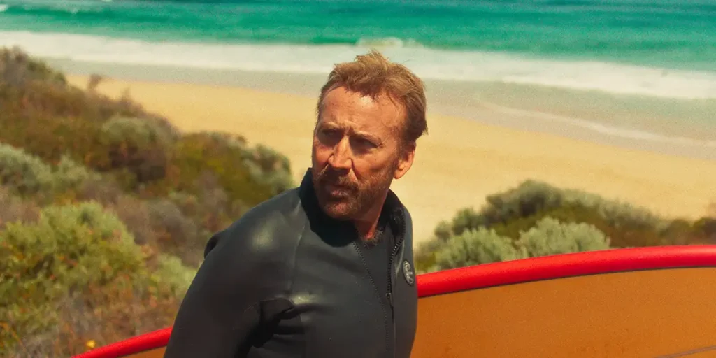 Nicholas Cage walks by the beach holding a surfboard in a still from The Surfer