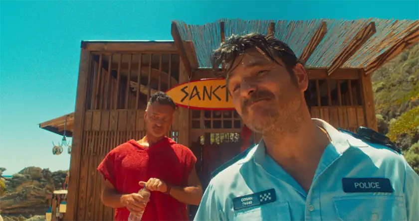 Two men stand outside a wooden building on the beach in a still from The Surfer