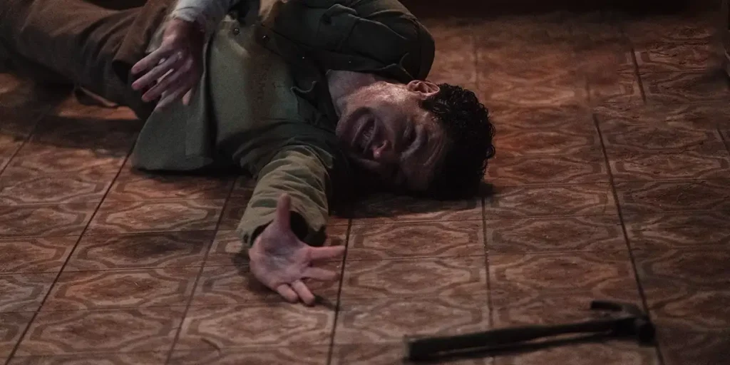 A man tries to reach a hammer while lying on the floor in pain in a still from the movie Wolf Man, used in an article about everything we know about the film