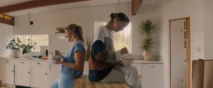 Two women lean on a wooden table in a kitchen eating lunch and smiling in a still from the film American Meltdown 