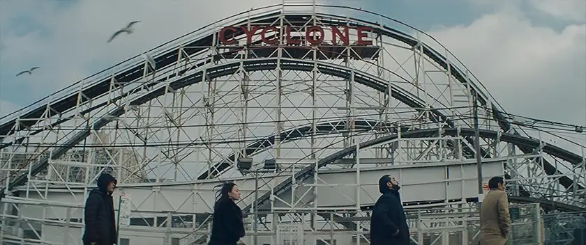 Yura Borisov, Mikey Madison, Vache Tovmasyan and Karren Karagulian walk in a row underneath a "Cyclone" rollercoaster in a still from the movie Anora, whose final scene and ending are explained in this article