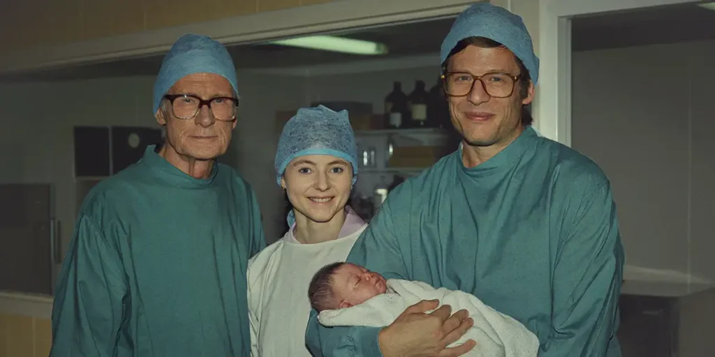 Bill Nighy, Thomasin McKenzie and James Norton wear hospital gowns and hold a newborn baby in a still from the film Joy