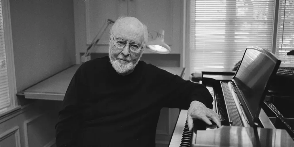 John Williams smiles, a hand placed on his piano, in a black and white still from the documentary film Music by John Williams