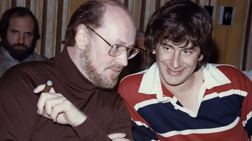 John Williams and Steven Spielberg have a conversation, the former smoking a cigar and the latter wearing a striped t-shirt and smiling, in the documentary film Music by John Williams