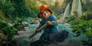 Paddington bear wears a raincoat while using an upside down umbrella as a boat on a river in a still from the 2024 film Paddington in Peru