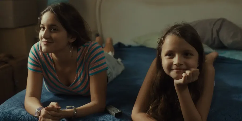 Two girls lie on a bed posing for a picture smiling in a still from the film Queens (Reinas)