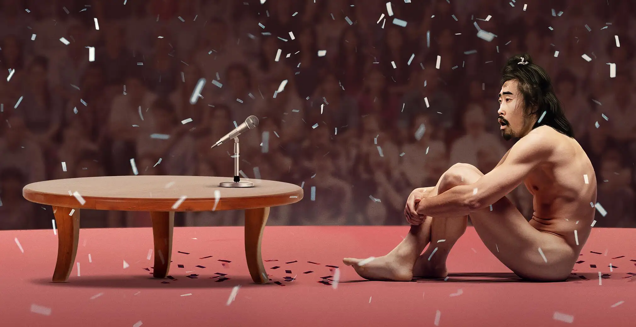 A naked man sits on the floor cross-legged in front of a table with a microphone on it, with confetti all around him, in the official Key Art for Hulu film The Contestant