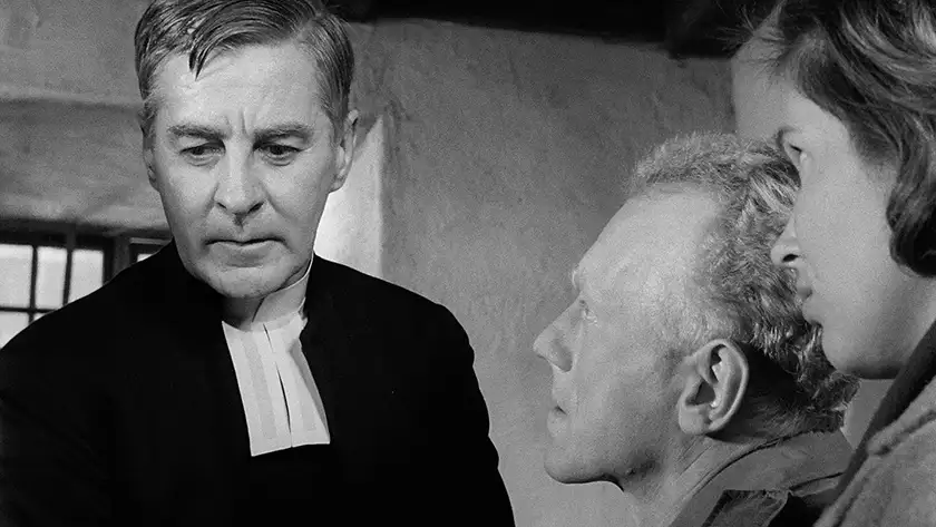 A priest looks to his right with two people looking at him in a still from the film Winter Light