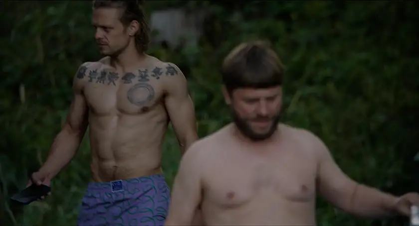 Two shirtless men walk in the woods in a still from the film Drowning Dry