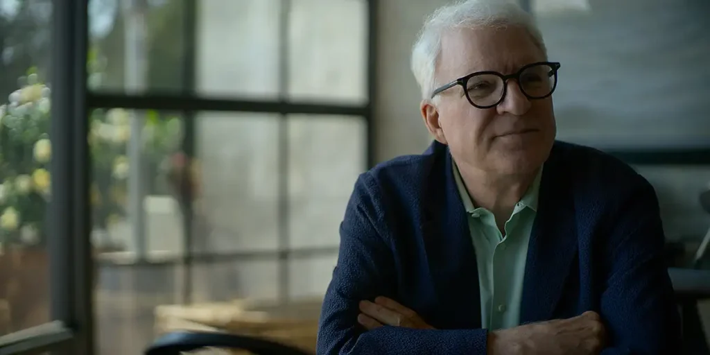 Steve Martin smiles looking to his left and leaning on a table in Apple TV+'s "STEVE! (martin) a documentary in 2 pieces"