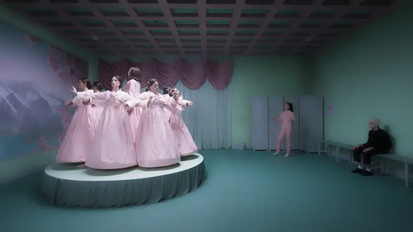 A group of young women wearing pastel pink nightgowns stands in a circle on a round stage looking outward in a green room, with another person dressed in pink in a corner and a boy wearing black sitting in front of them, in a still from the Eduardo Casanova film La Pietà