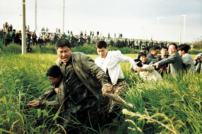 Song Kang Ho and Kim Sang-Kyung are in a corn field with a lot of other people, all hanging on to someone, in Memories of Murder