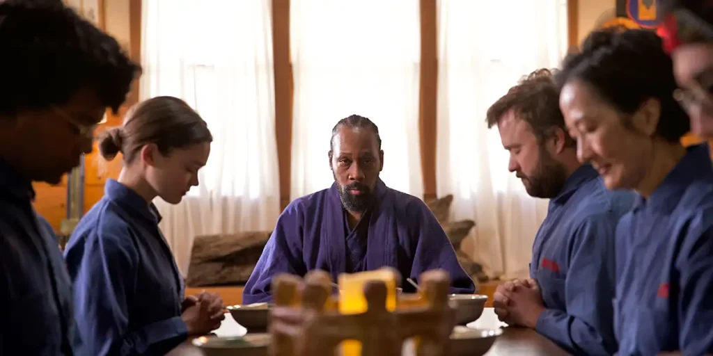 RZA sits at a dinner table looking ahead with people all around the table wearing blue robes in a still from the film Not An Artist