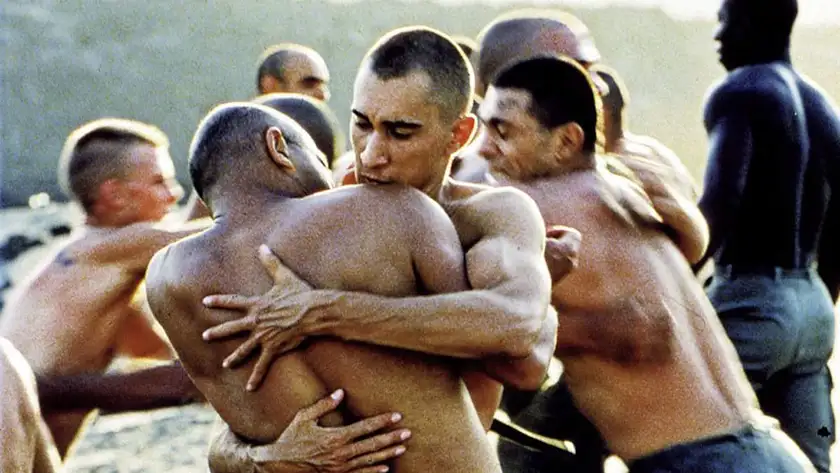 Two shirtless soldiers hug each other outdoors with more shirtless men behind them in a scene from Beau Travail