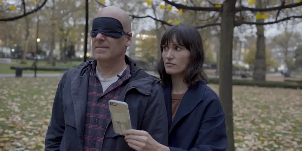 A woman holds a phone and accompanies a blindfolded man through a park in the film My Husband, The Cyborg