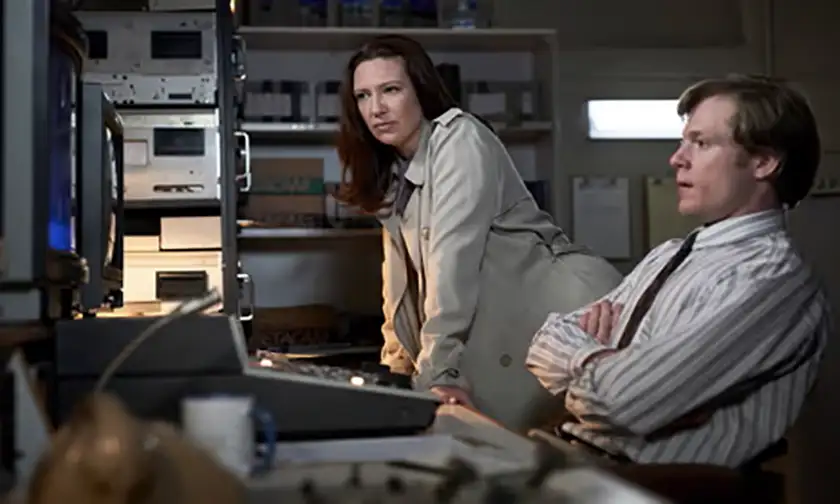 A man and a woman sit looking at the screens of a live show in a television studio in a still from the series The Newsreader