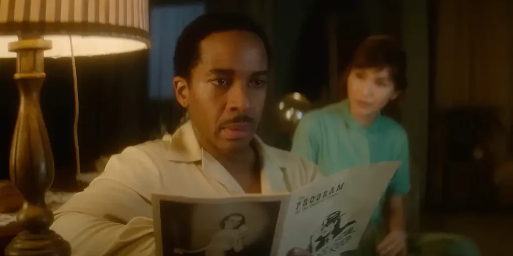 A man sits on the couch looking up with an open leaflet in his hands, with a woman sitting behind him looking at him, in a still from the 2025 movie The Actor