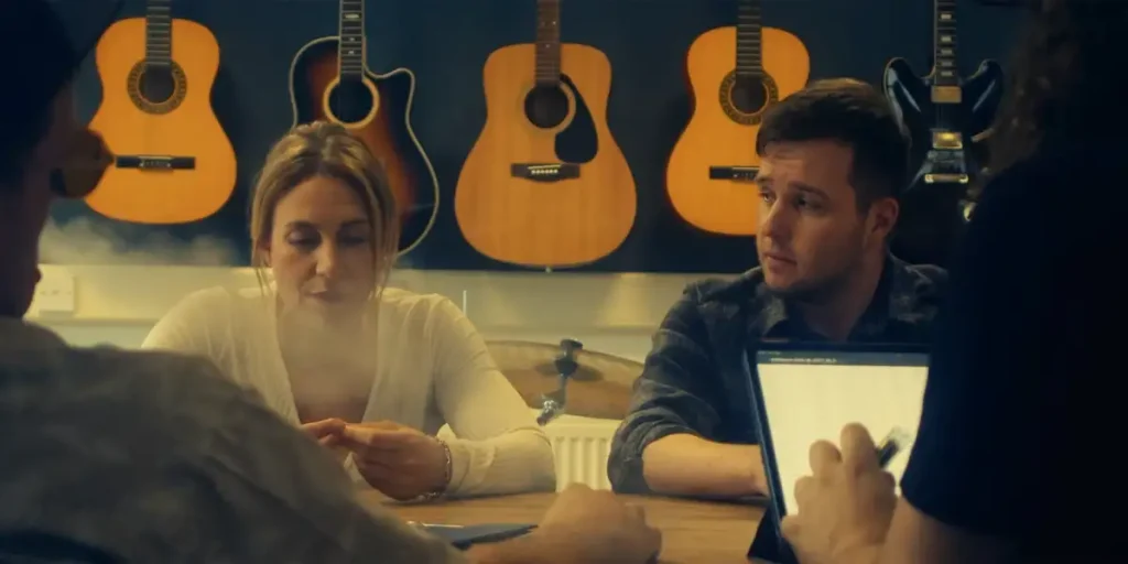A man and a woman sit at a table with some guitars behind them in a still from the 2025 film "Kid"