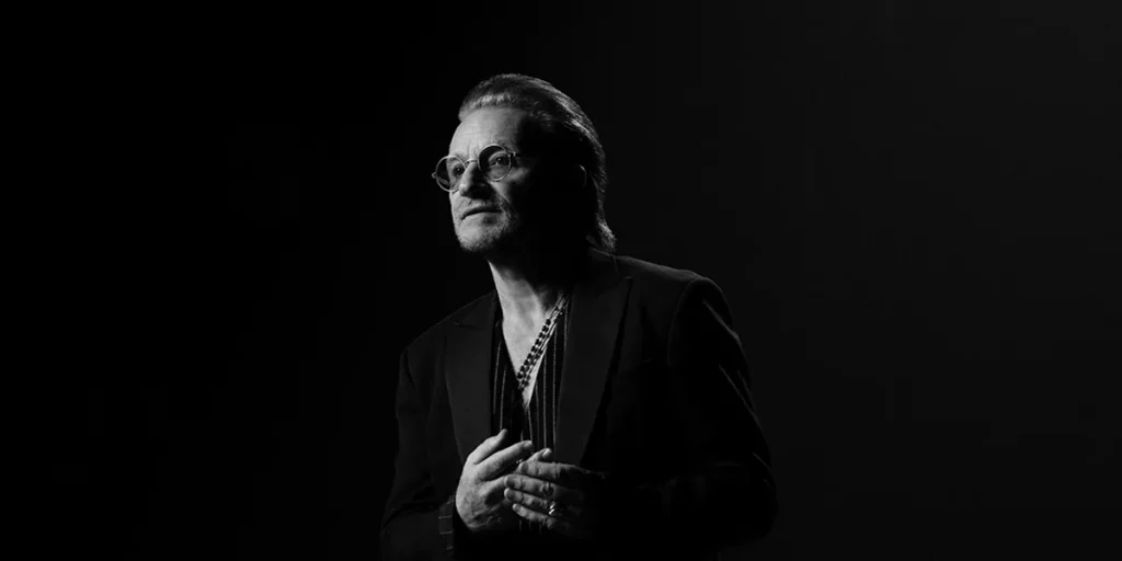 Bono has his hands on his chest in a black and white still from Bono: Stories of Surrender