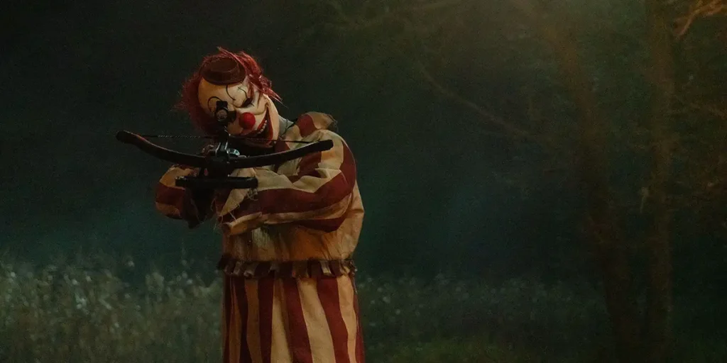 A clown holds a bow and arrow in a still from Clown in a Cornfield