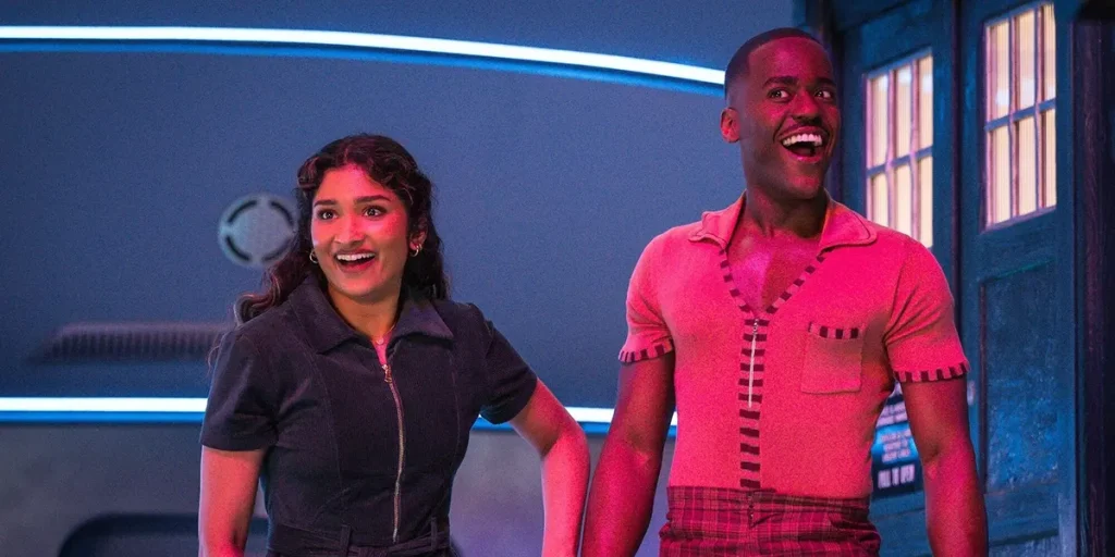 Varada Sethu and Ncuti Gatwa, as The Doctor and Belinda, look excited in a still from Season 15 (also known as Season 2) Episode 6 of Doctor Who (2025), titled "The Interstellar Song Contest"