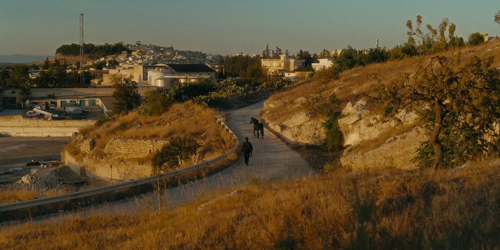 A man and a horse walk up a road in between hills in a still from the film Siticulosa