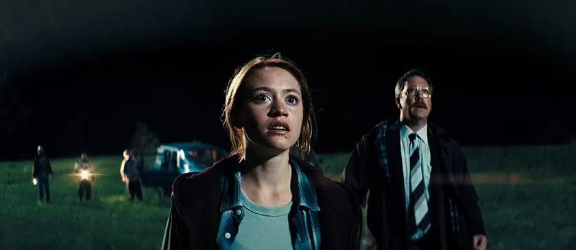 A young woman looks at the sky at night, with a man with a tie behind her, in a still from the movie Watch the Skies