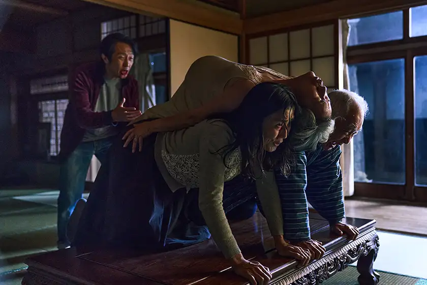 A man and a woman sit on a table with another woman on top of them facing the ceiling and a man looking at them in despair in a still from the horror movie Best Wishes to All (2025)