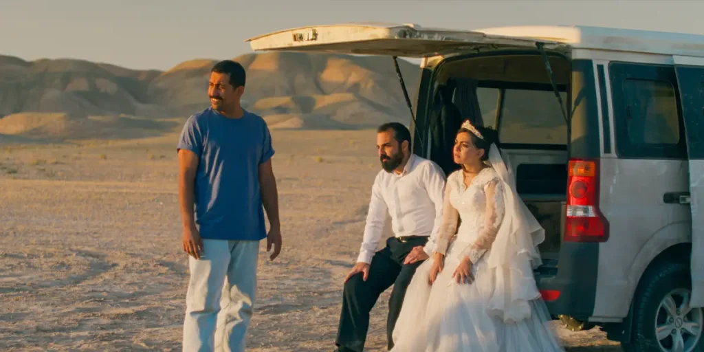 A woman wears a wedding dress and sits next to a man, with another man standing next to them, by a car in a still from the Jafar Panahi movie It Was Just an Accident (Yek Tasadef Sadeh) featured in Loud and Clear Reviews' review of the film