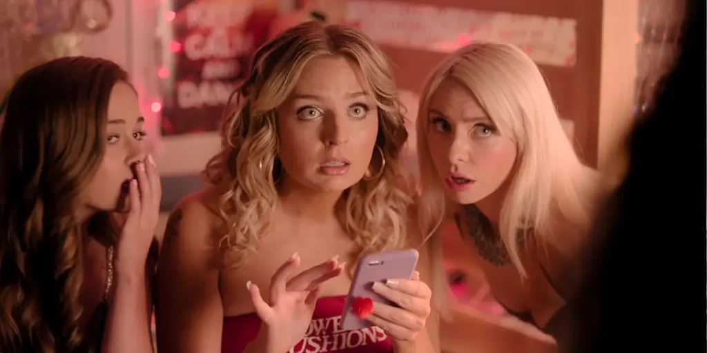 Three girls look up after looking at a phone screen, which one of them is holding, in a still from Overcompensating Season 1