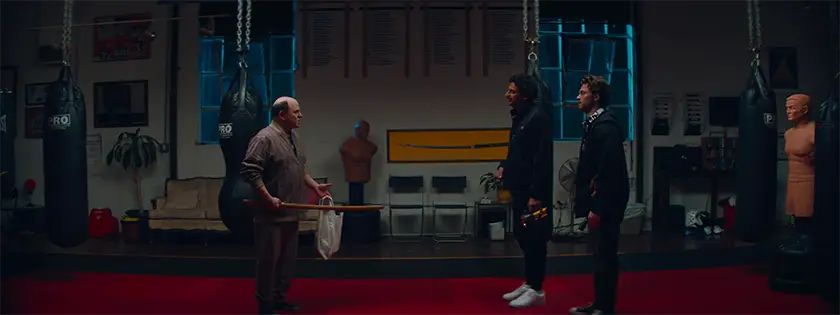 A man holds a katana in a gym, facing two other men, in a scene from Stealing Pulp Fiction