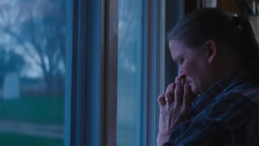 Victoria Lopez cries with her eyes closed and her hands in preyer, standing facing a window, in the short film "Watch Over Us" (2025)