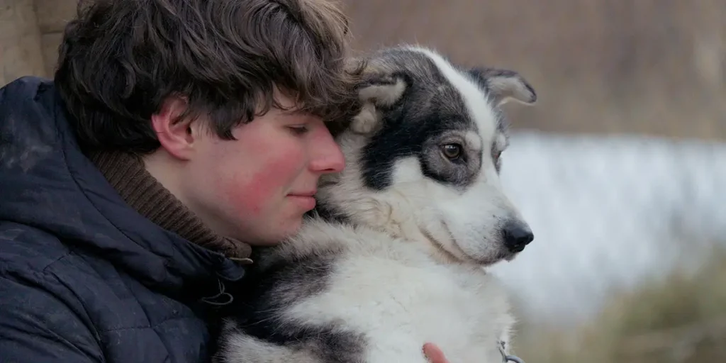 A young man named Romain hugs a dog named Mjød, outdoors, in the 2025 documentary movie Folktales, from directors Heidi Ewing and Rachel Grady
