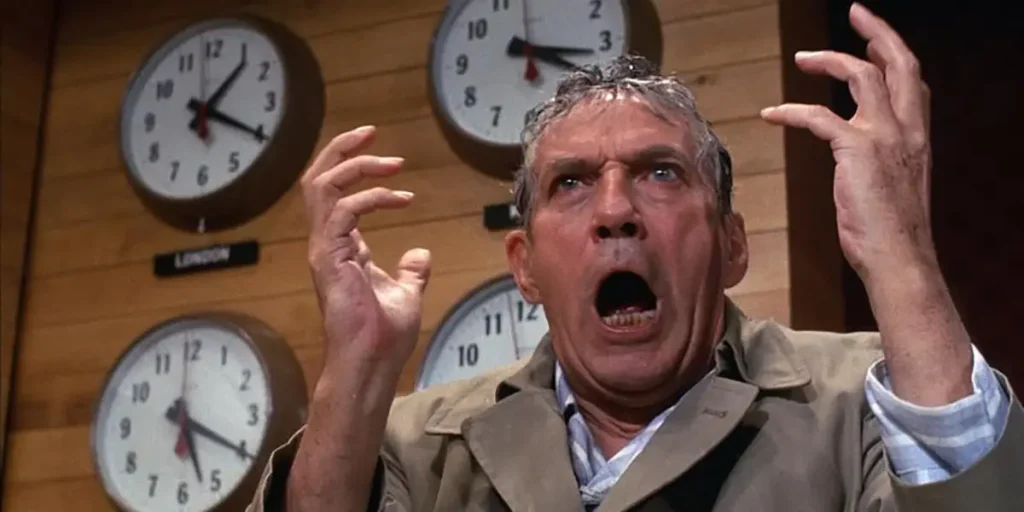 Peter Finch stands with arms open in shock in front of some clocks in a still from the movie Network (1976)