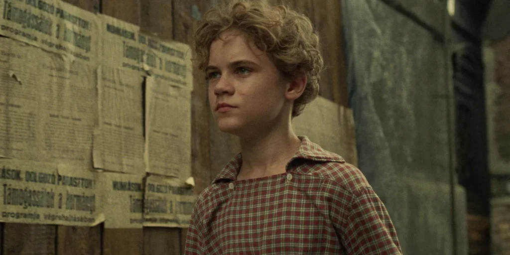 A young man stands next to a wall with some newspaper pages on it in the film Orphan (Árva), from director László Nemes