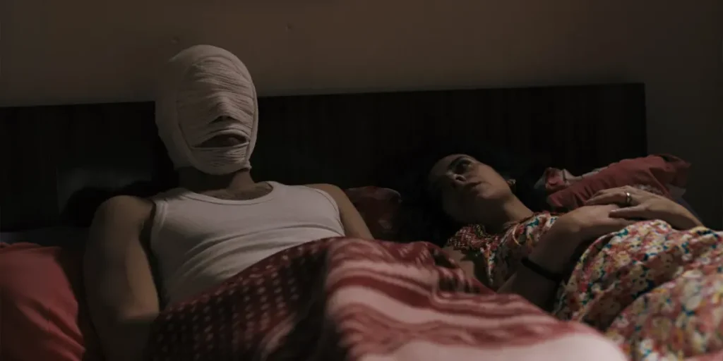A man with a bandaged head and a woman lie in bed in a still from Roqia
