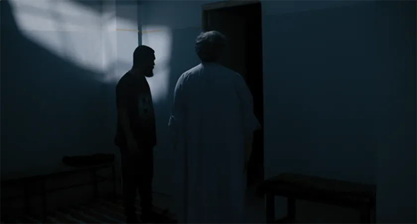Two men stand in a dark room in a still from Roqia
